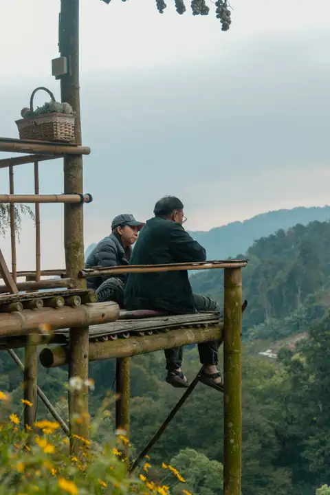 Two people seated on a bamboo platform overlooking forested hills beneath a cloudy sky