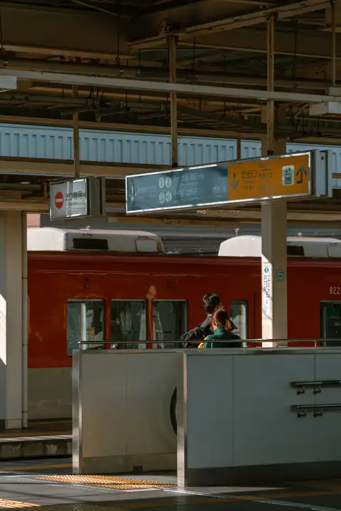 A red commuter train stopped behind the concrete structures of a sunlit station platform