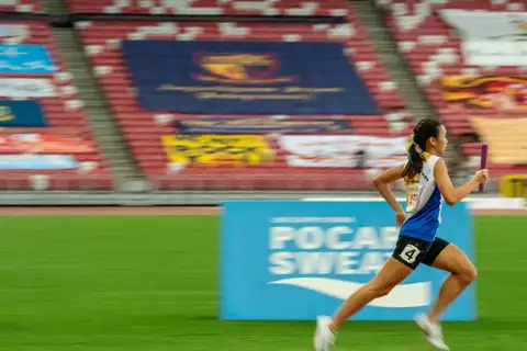 A student relay runner carrying a purple baton at full stride against a motion-blurred stadium